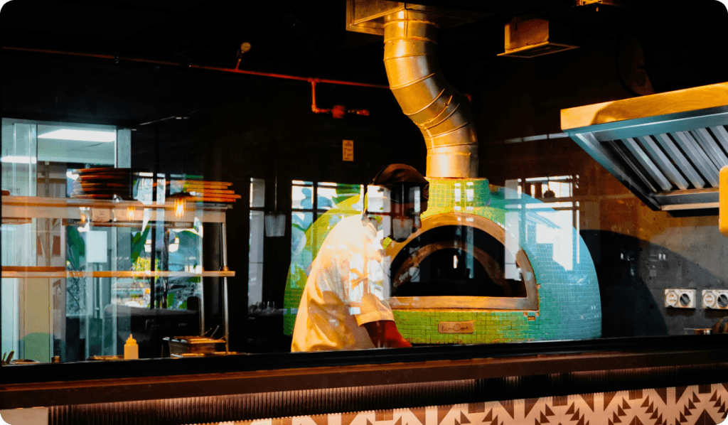 Chef working at a green-tiled wood-fired pizza oven in an open restaurant kitchen, viewed through a glass partition